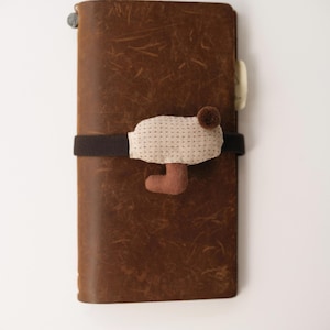 May include: A dark brown leather notebook secured with a dark brown elastic band. Attached to the band is a small, plush, beige fabric mushroom ornament with a brown stem and a brown dot on the cap, featuring a subtle grid pattern.