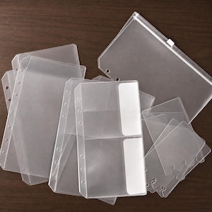 May include: A collection of clear plastic binder inserts and pouches. The set includes multiple rectangular sheets with punched holes along the side, a zippered pouch, and smaller card-sized pockets with white opaque sections for labeling.