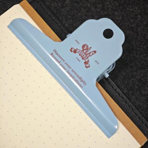 May include: A light blue metal binder clip with a scalloped top edge and a circular hole. The clip features a brown logo with a magnifying glass and quill, plus the text: Discover your serendipity, Record your inspirations, ESQUARE STUDIO 2016.