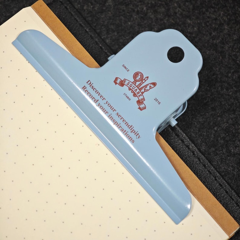 May include: A light blue metal binder clip with a scalloped top edge and a circular hole. The clip features a brown logo with a magnifying glass and quill, plus the text: Discover your serendipity, Record your inspirations, ESQUARE STUDIO 2016.