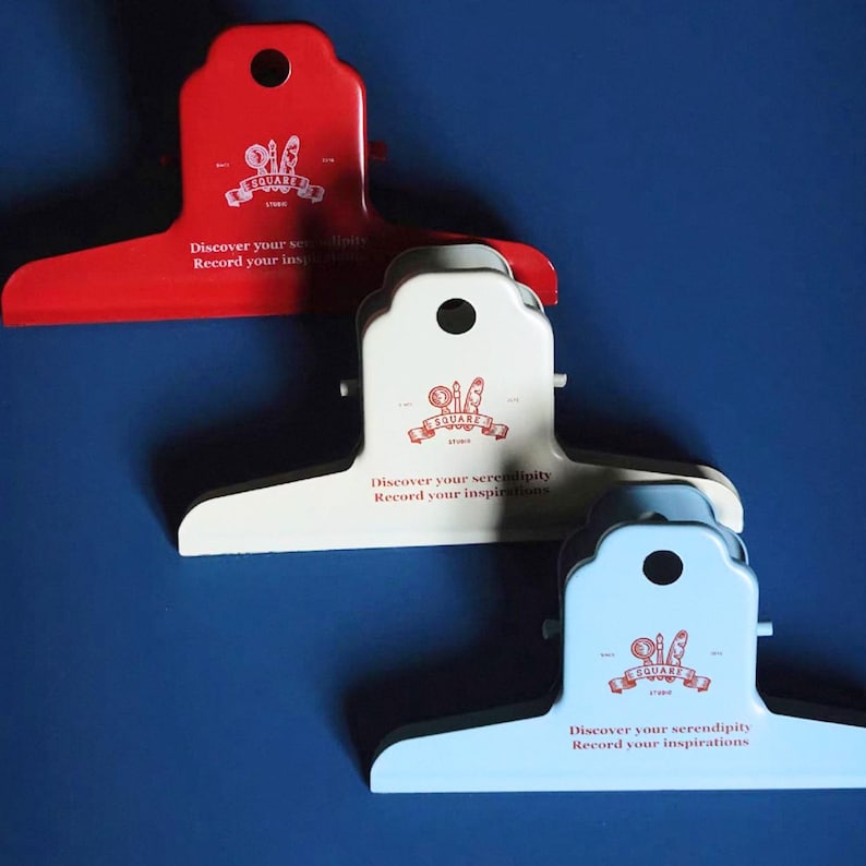 May include: Three metal bulldog clips in red, cream, and light blue. Each clip features a circular hole at the top and a printed logo with the text "SQUARE STUDIO" above the phrase "Discover your serendipity Record your inspirations" in red ink.