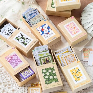 Könnte beinhalten: Mehrere rechteckige Pappschachteln mit Stapeln kleiner, perforierter, briefmarkenförmiger Papierkarten. Jede Schachtel zeigt auf der Vorderseite eine einzigartige Blumen- oder Pflanzenillustration mit gedruckten Textetiketten wie Iris Letter, Twilight Messenger und Sunlit Floral Mail.