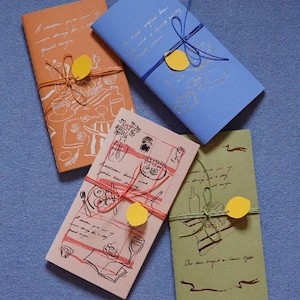 Puede incluir: Cuatro cuadernos rectangulares en naranja, azul, rosa y verde, cada uno con ilustraciones dibujadas a mano de comida y bebida, texto cursivo, un cordel de color y un pequeño recorte de limón de papel amarillo en la portada.