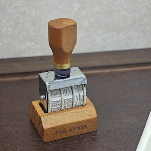 May include: A vintage-style date stamp featuring a wooden handle and a chrome-plated metal frame. The device includes rotating white plastic wheels for adjusting the date and month. The wooden base is engraved with the text PAR-AVION in dark ink.