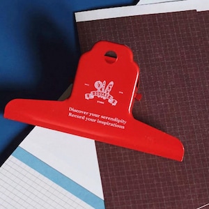 May include: A bright red metal bulldog clip featuring a white logo with a magnifying glass, pen, and brush, along with the printed text: Discover your serendipity, Record your inspirations. The clip has a circular hole at the top for hanging.