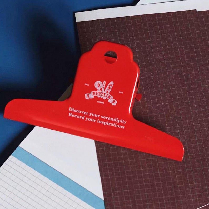May include: A bright red metal bulldog clip featuring a white logo with a magnifying glass, pen, and brush, along with the printed text: Discover your serendipity, Record your inspirations. The clip has a circular hole at the top for hanging.