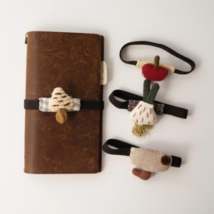 May include: A brown leather notebook secured with a black elastic band featuring a plush mushroom charm. Three additional black elastic bands are included, each adorned with fabric charms shaped like a red apple, a white garlic bulb, and a beige pouch.