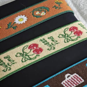 May include: Three strips of woven fabric trim featuring embroidered designs. The top strip is rust-colored with white daisies and green foliage. The middle strip is beige with red cherries and the text Sweet Time. The bottom strip is brown with white baskets.