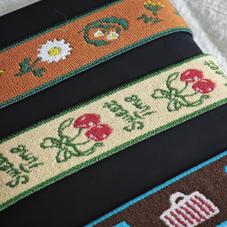 May include: Three strips of woven fabric trim featuring embroidered designs. The top strip is rust-colored with white daisies and green foliage. The middle strip is beige with red cherries and the text Sweet Time. The bottom strip is brown with white baskets.