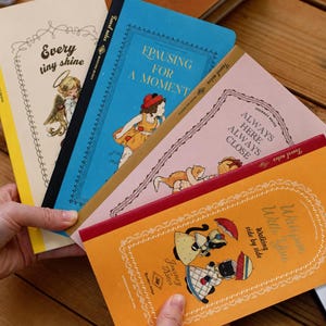 May include: Four rectangular notebooks with illustrated covers. The cream notebook says Every tiny shine, the blue one says PAUSING FOR A MOMENT, the pink one says ALWAYS HERE ALWAYS CLOSE, and the orange one says Walking side by side.