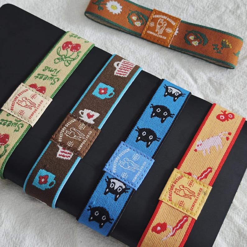 May include: Five elastic bands featuring woven patterns including cherries, mugs, black cats, and animals. Each band has a rectangular fabric patch with the text BENSTORE DESIGN EST 2013 GENPOO.LTD.CO and a graphic of two standing cats.