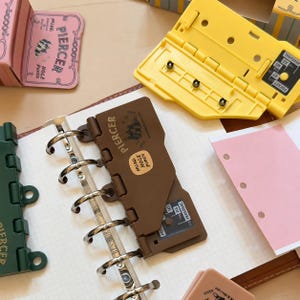 May include: A brown plastic mini hole punch tool with the text PIERCER and MINI HOLE PUNCH printed on the surface. The tool features a metal ring mechanism and is positioned on a grid paper sheet. Additional yellow and green hole punch tools are visible.