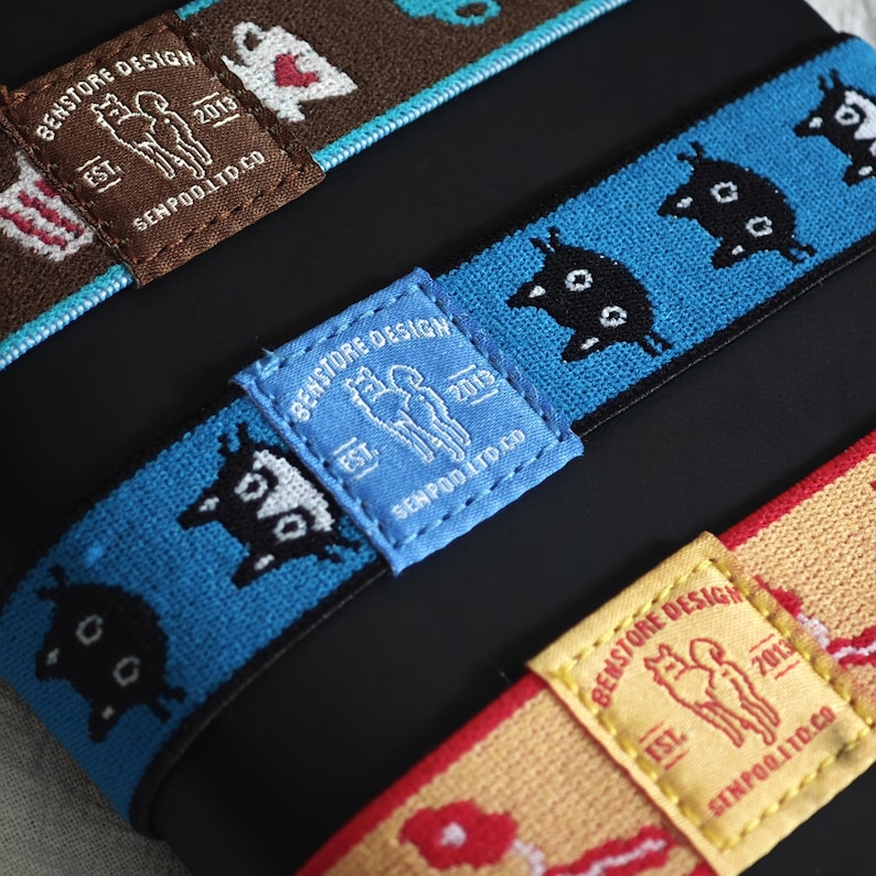 May include: Three elastic bands featuring woven patterns and rectangular fabric labels. The labels display the text BENSTORE DESIGN EST. 2013 SENPOD.LTD.CO with a graphic of two figures. The bands are brown, blue, and yellow with various icons.