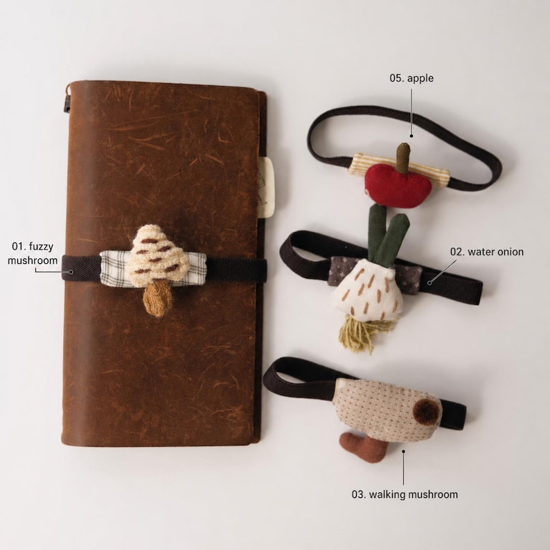 May include: A brown leather notebook with a fuzzy mushroom elastic band, alongside three elastic bands featuring fabric charms: a red apple, a white water onion, and a beige walking mushroom, each labeled with numbers and names.