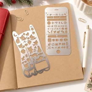 May include: Two silver metal stencils for journaling and planning. One stencil is shaped like a cat and features cutouts of cats, hearts, stars, and music notes. The other rectangular stencil features icons for phone interfaces, social media, and navigation.