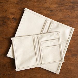 May include: Two off-white fabric wallets featuring a bifold design with multiple interior card slots and a main compartment. The wallets are constructed from a durable, plain-weave textile with reinforced stitched edges along the perimeter.