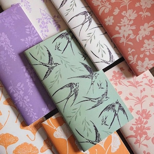 May include: A collection of rectangular paper sheets featuring various patterns including black swallows on sage green, white floral prints on coral, purple wisteria-like designs, and white ginkgo leaves on orange and white backgrounds.
