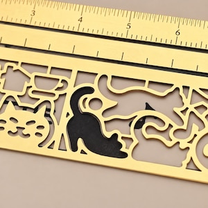 Könnte beinhalten: Ein goldfarbenes Metall-Schablonenlineal mit ausgeschnittenen Motiven von Katzen, einem Haus, einer Teetasse und Musiknoten. Das Lineal zeigt imperiale Maßeinheiten, wobei der sichtbare Bereich Messungen zwischen 3 und 6 Zoll am oberen Rand aufweist.