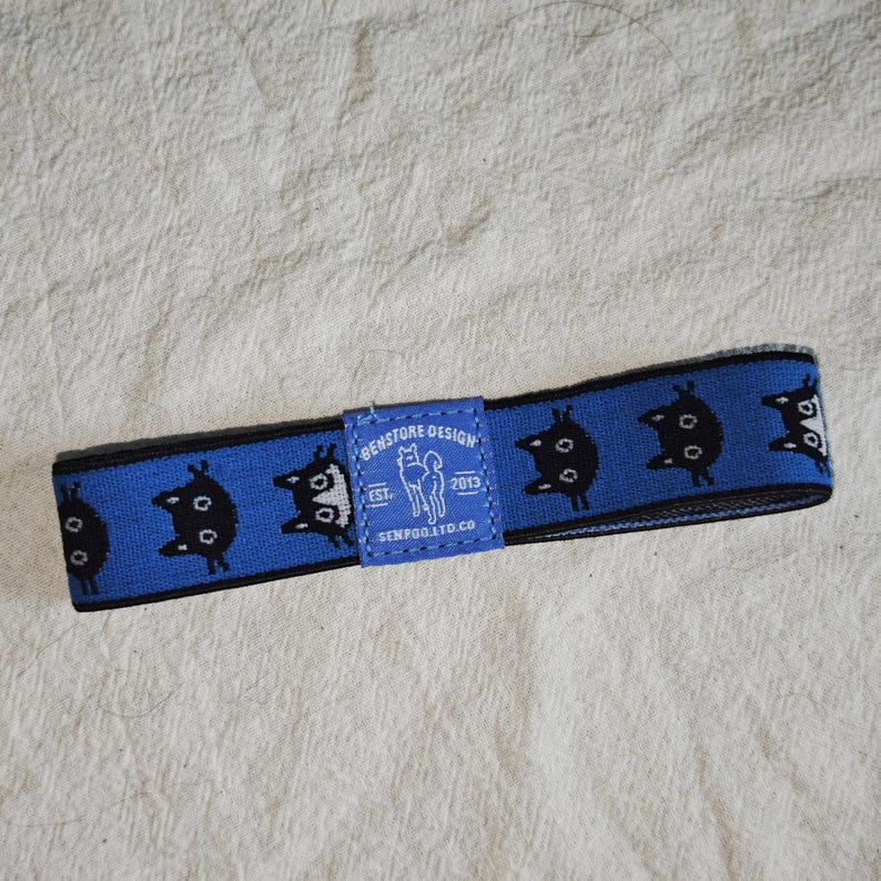May include: A blue elastic headband featuring a repeating pattern of black cat silhouettes. A central blue fabric label displays the text "BENSTORE DESIGN EST. 2013 SENPOO.LTD.CO" along with a small white graphic of a dog standing in profile.