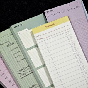 May include: Three stacked paper notepads with different layouts. The top pad is yellow with a TO DO LIST header, date and subject lines, and a checklist. The middle pad is sage green labelled FOCUS ON with lined sections. The back pad is lavender with a TIMELINE header and grid.