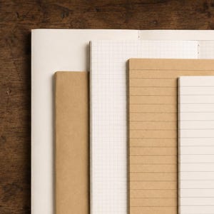 May include: A collection of five notebooks arranged in a layered stack. The set includes notebooks with plain white pages, white grid paper, and tan covers with horizontal lined pages, showcasing various paper styles and textures for writing.