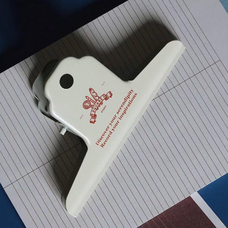 May include: A white metal binder clip featuring a red logo with the text SQUARE STUDIO and the phrase Discover your serendipity Record your inspirations printed in red on the front surface. The clip has a circular cutout on the top handle.