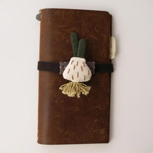 May include: A rectangular brown leather notebook secured with a black elastic band. Attached to the band is a fabric ornament shaped like a white bulb with green stems and a light green fringed base, featuring small brown embroidered dash details.