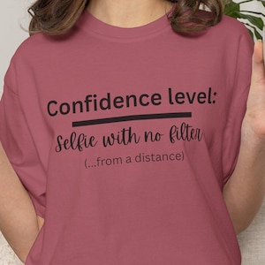 Puede incluir: Una camiseta malva con el texto "Confidence level: Selfie with no filter (...from a distance)". La persona lleva pantalones cortos de mezclilla y sostiene una taza blanca con borde marrón. La persona también hace el signo de la paz.