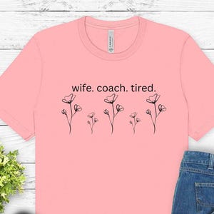 May include: A light pink t-shirt with the text "wife. coach. tired." and a floral design. The shirt is paired with blue jeans and black canvas sneakers with white laces.