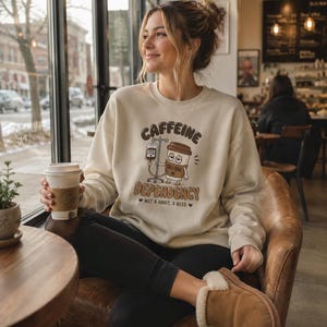 May include: A cream-colored crewneck sweatshirt featuring a graphic of a coffee cup connected to an IV drip. The text reads CAFFEINE DEPENDENCY and NOT A HABIT, A NEED. The design includes brown and black lettering with a cartoon coffee cup.