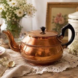 May include: A copper tea kettle with a black handle and lid knob sits on a lace doily. A teacup and saucer with floral designs are in the foreground. A vase of white flowers and a framed picture are in the background.
