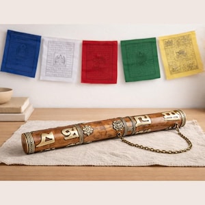 May include: A wooden prayer wheel with gold-colored accents and a chain, resting on a beige cloth. Colorful prayer flags hang in the background. The prayer wheel is brown with gold-colored metal bands and decorative elements.