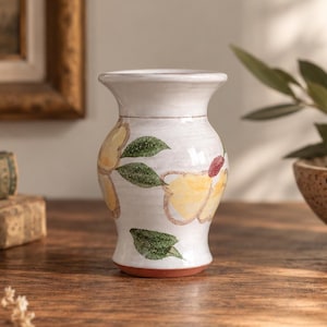 May include: A white ceramic vase with a flared rim and a tapered base, featuring a hand-painted floral design with yellow petals, green leaves, and small red accents. The vase has a smooth, glossy finish and a visible terracotta-colored base rim.