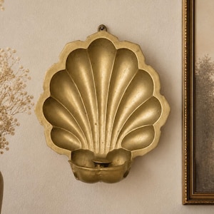 May include: A gold-colored, shell-shaped wall sconce with a small, round base for holding a candle. The sconce is mounted on a neutral-colored wall. A framed picture and a vase with dried flowers are visible in the background.