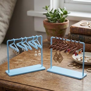 May include: Two light blue metal jewelry display stands, each featuring a rectangular base and a horizontal bar. One stand holds light blue miniature hangers with gold hoop and pearl earrings, while the other holds brown miniature hangers with dangling earrings.