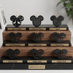 May include: A three-tiered display stand holding nine black mouse-shaped earrings. Each pair is labeled with gold plaques reading Tree of Life, Chevron Wave, Swirl, Art Nouveau, Linear Art, Floral Bow, Spiral Web, Polka Dots, Globe Grid, and Magical Earring Collection.