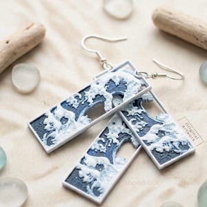May include: A pair of rectangular drop earrings featuring a raised, textured design of white crashing waves against a dark blue background, framed in white. The earrings include silver-toned metal hooks and a small label reading Original Artwork.