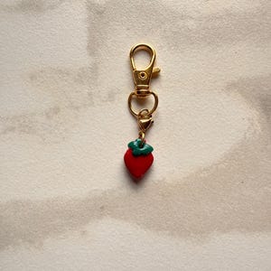 K&ouml;nnte beinhalten: Ein goldfarbener Metall-Karabiner-Schl&uuml;sselanh&auml;nger mit einem kleinen, roten Erdbeer-Anh&auml;nger mit gr&uuml;nem Stiel. Der Erdbeer-Anh&auml;nger ist &uuml;ber einen kleineren goldenen Biegering und einen zweiten Karabinerverschluss mit dem Hauptkarabiner verbunden.