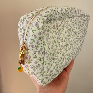 K&ouml;nnte beinhalten: Ein gestepptes T&auml;schchen mit wei&szlig;em Stoffgrund und einem kleinen violetten Blumenmuster mit gr&uuml;nen Bl&auml;ttern. Das T&auml;schchen hat einen goldfarbenen Rei&szlig;verschluss mit einem kleinen gelben Fruchtanh&auml;nger und eine rautenf&ouml;rmige Steppung.