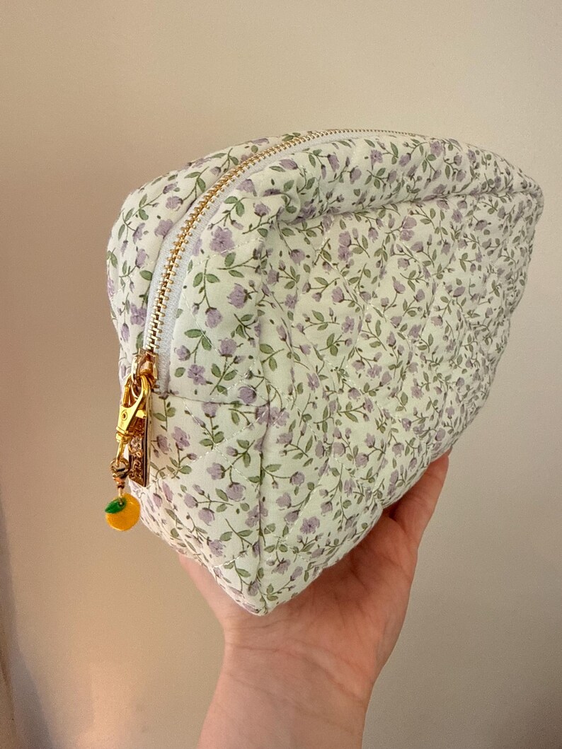 K&ouml;nnte beinhalten: Ein gestepptes T&auml;schchen mit wei&szlig;em Stoffgrund und einem kleinen violetten Blumenmuster mit gr&uuml;nen Bl&auml;ttern. Das T&auml;schchen hat einen goldfarbenen Rei&szlig;verschluss mit einem kleinen gelben Fruchtanh&auml;nger und eine rautenf&ouml;rmige Steppung.