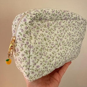 K&ouml;nnte beinhalten: Ein gestepptes T&auml;schchen mit wei&szlig;em Stoffgrund und einem sich wiederholenden Muster aus kleinen violetten Blumen und gr&uuml;nen Bl&auml;ttern. Das T&auml;schchen hat einen goldfarbenen Rei&szlig;verschluss mit einem kleinen Anh&auml;nger in Form einer gelben Zitrone.