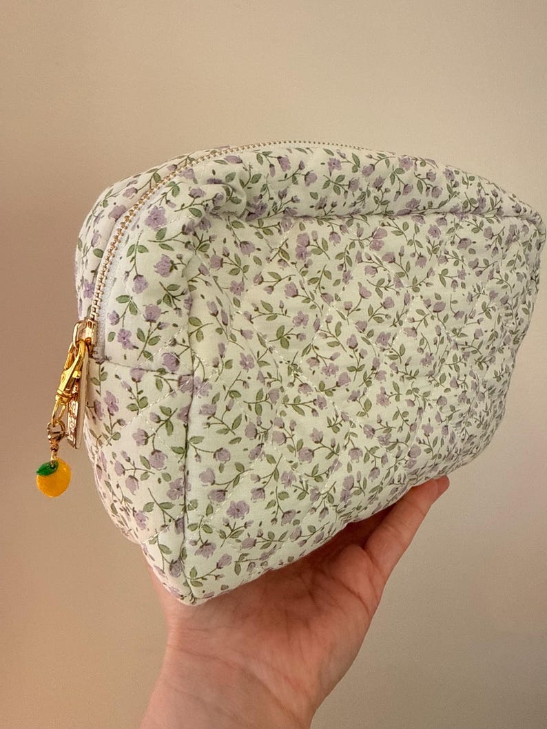 K&ouml;nnte beinhalten: Ein gestepptes T&auml;schchen mit wei&szlig;em Stoffgrund und einem sich wiederholenden Muster aus kleinen violetten Blumen und gr&uuml;nen Bl&auml;ttern. Das T&auml;schchen hat einen goldfarbenen Rei&szlig;verschluss mit einem kleinen Anh&auml;nger in Form einer gelben Zitrone.