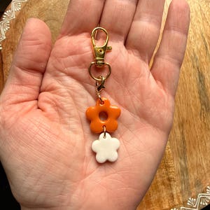 K&ouml;nnte beinhalten: Ein Schl&uuml;sselanh&auml;nger aus goldfarbenem Metall mit Karabinerverschluss und zwei &uuml;bereinander angeordneten Blumenanh&auml;ngern. Die obere Blume ist gl&auml;nzend orange, die untere Blume ist gl&auml;nzend wei&szlig;, verbunden durch kleine goldfarbene Metallringe.