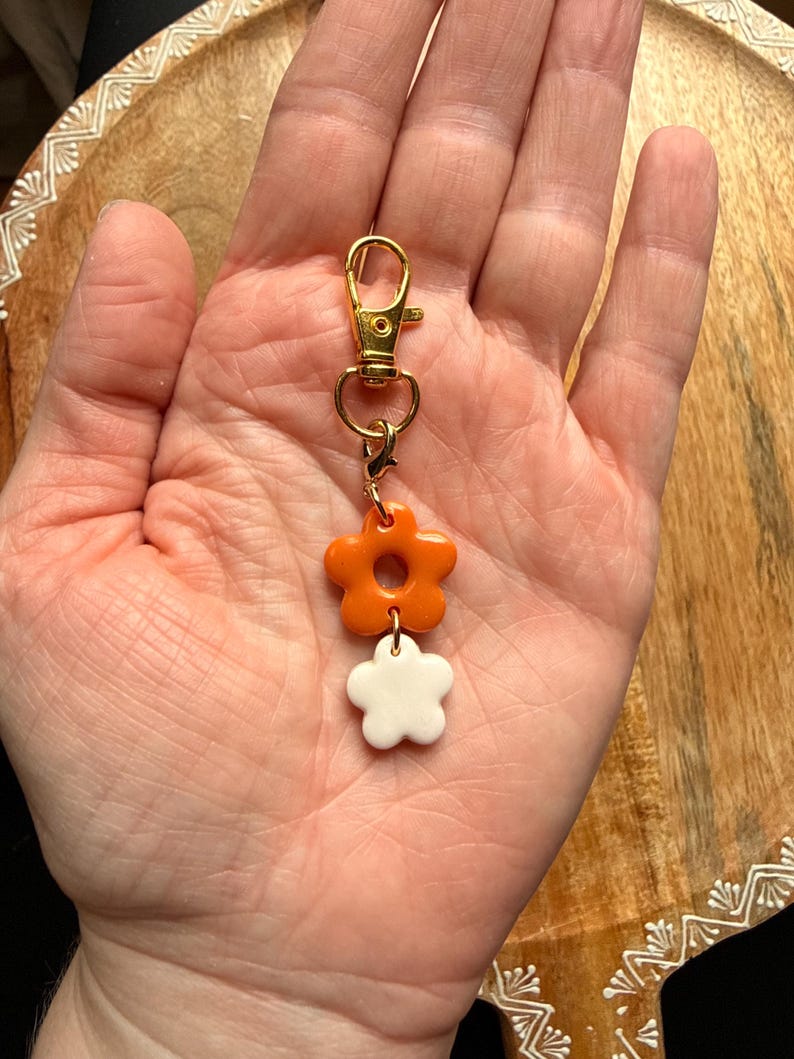 K&ouml;nnte beinhalten: Ein Schl&uuml;sselanh&auml;nger aus goldfarbenem Metall mit Karabinerverschluss und zwei &uuml;bereinander angeordneten Blumenanh&auml;ngern. Die obere Blume ist gl&auml;nzend orange, die untere Blume ist gl&auml;nzend wei&szlig;, verbunden durch kleine goldfarbene Metallringe.