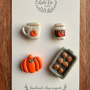 May include: A set of four handmade clay magnets on a display card labeled Lala Vie & Co. The magnets include a white mug with a pumpkin, a white coffee cup with a pumpkin, an orange pumpkin, and a gray baking tray with six brown muffins.