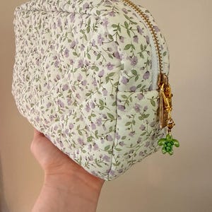 K&ouml;nnte beinhalten: Ein gestepptes T&auml;schchen mit wei&szlig;em Stoffgrund und einem sich wiederholenden Muster aus kleinen violetten Blumen und gr&uuml;nen Stielen. Das T&auml;schchen hat einen goldfarbenen Rei&szlig;verschluss mit einem kleinen gr&uuml;nen Schildkr&ouml;tenanh&auml;nger am Schieber.