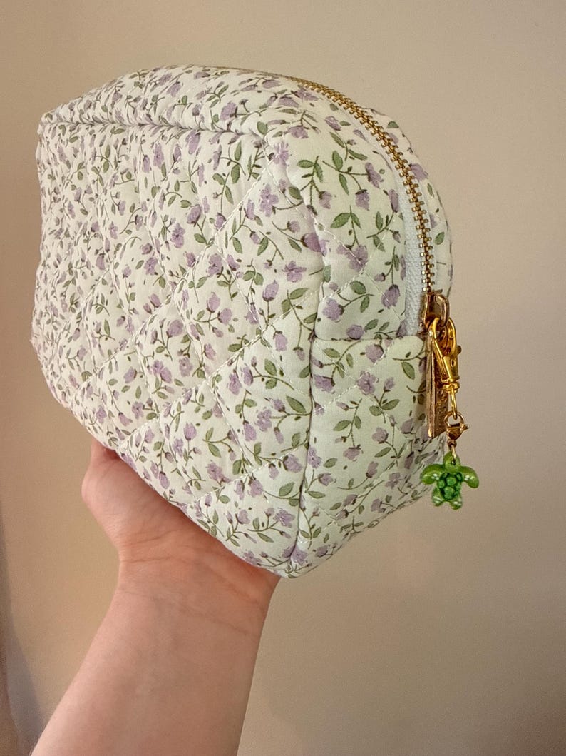 K&ouml;nnte beinhalten: Ein gestepptes T&auml;schchen mit wei&szlig;em Stoffgrund und einem sich wiederholenden Muster aus kleinen violetten Blumen und gr&uuml;nen Stielen. Das T&auml;schchen hat einen goldfarbenen Rei&szlig;verschluss mit einem kleinen gr&uuml;nen Schildkr&ouml;tenanh&auml;nger am Schieber.