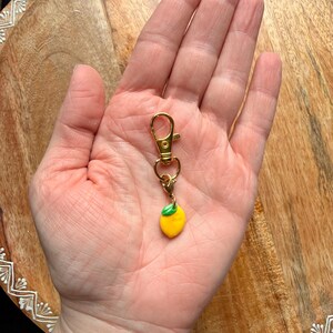 K&ouml;nnte beinhalten: Ein Schl&uuml;sselanh&auml;nger mit Karabinerverschluss aus goldfarbenem Metall, der einen kleinen, glatten gelben Zitronenanh&auml;nger mit einem einzelnen gr&uuml;nen Blatt an der Oberseite aufweist. Der Anh&auml;nger h&auml;ngt an einem Ring, der mit dem Karabiner verbunden ist.