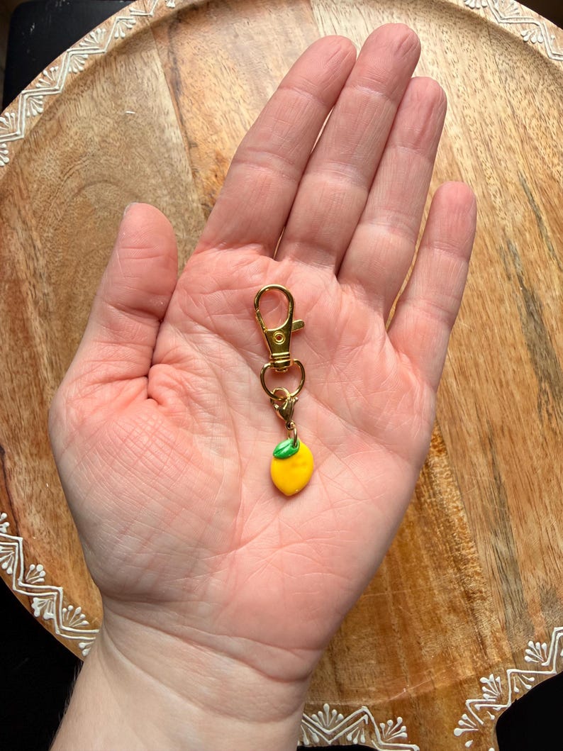 K&ouml;nnte beinhalten: Ein Schl&uuml;sselanh&auml;nger mit Karabinerverschluss aus goldfarbenem Metall, der einen kleinen, glatten gelben Zitronenanh&auml;nger mit einem einzelnen gr&uuml;nen Blatt an der Oberseite aufweist. Der Anh&auml;nger h&auml;ngt an einem Ring, der mit dem Karabiner verbunden ist.