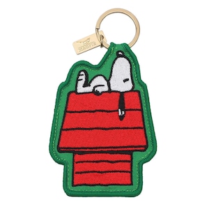 May include: A keychain featuring an embroidered patch of a white dog with black ears lying on a red doghouse. The patch has a green border and is attached to a gold-toned metal ring with a small rectangular tag engraved with the word MAGPIE.